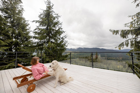 Woman in pink sweater rests with her dog on a spacious white terrace in mountains. Recreation in house on nature conceptの写真素材