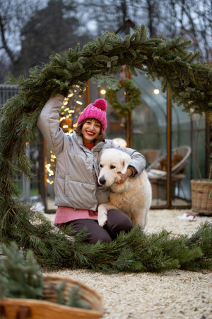 Portrait of a cute woman with her dog hugs together in a big Christmas wreath at backyard. Concept...