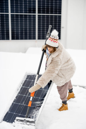 Woman cleans solar panels from snow to produce power in winter on the roof of her house. Energy...