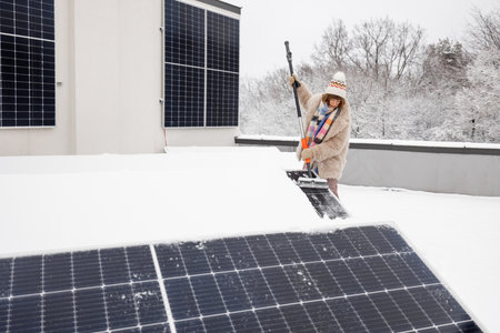 Woman cleans solar panels from snow to produce power in winter on the roof of her house. Energy independence and sustainability conceptの写真素材