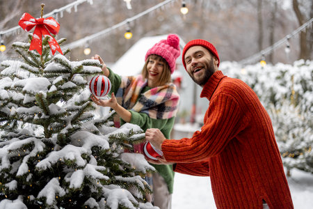 Man and woman decorate Christmas tree with festive balls, while preparing for a winter holidays at snowy backyard of their house. Happy family celebrating New Years holidaysの写真素材
