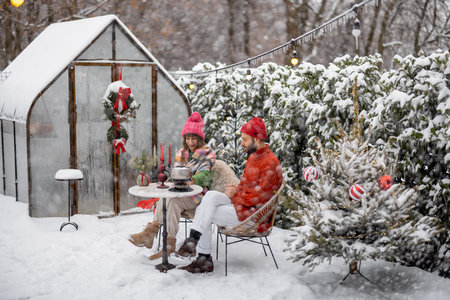 Man and woman have romantic dinner, while sitting together by the table at beautifully decorated snowy backyard. Young family celebrating winter holidaysの写真素材