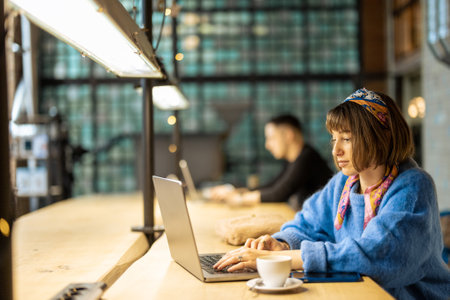Young stylish woman works on laptop while sitting by shared table with a coffee drink at modern cafe. Concept of remote work from public place, digital freelance and modern lifestyleの写真素材