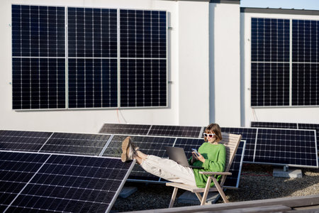 Woman works on laptop and phone while sitting on rooftop with a solar station. Concept of remote...