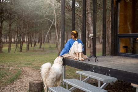 Young woman cares her dog while sitting on a porch of wooden house in pine forest, spending leisure time together and resting at countrysideの写真素材
