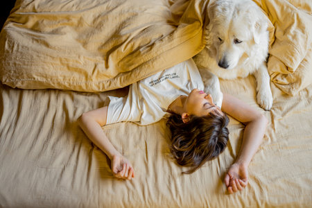 Young woman hugs with her cute dog while lying together covered with beige blanket in bed. View from above. Concept of friendship with pets and home cozinessの写真素材
