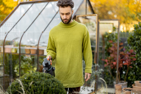 Stylish guy takes care of plants spraying on on them in garden with glasshouse on background....