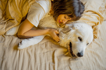 Young woman hugs with her cute dog while lying together covered with beige blanket in bed. Concept of friendship with pets and home cozinessの写真素材