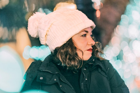 Curly-haired blonde woman with white cap looking to the side with Christmas lights on out-of-focus backgroundの写真素材