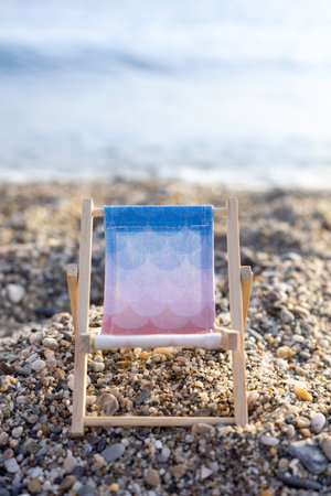 Close-up of a colorful miniature beach chair placed on a pebbled shoreline, with the sea softly blurred in the background. The bright light and centered composition evokes a playful, relaxed summer atmosphere.の写真素材