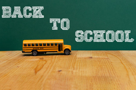 A yellow school bus with its doors open, on a wooden table under a green chalkboard displaying the...