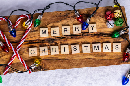 Merry Christmas spelled out using wooden letter tiles on a rustic wooden background; surrounded by colorful Christmas lights; candy canes; and festive jingle bellsの写真素材