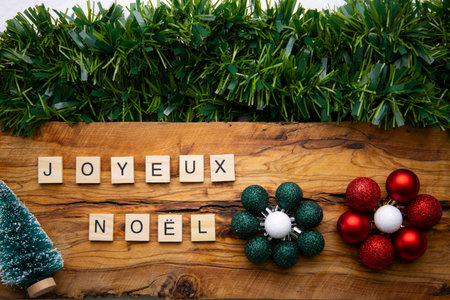 Festive image with the message âJoyeux NoÃ«lâ written with wooden tiles on a wooden base, accompanied by green garlands, a snow-covered pine tree, and spherical decorations in red, green, and white with glitterの写真素材