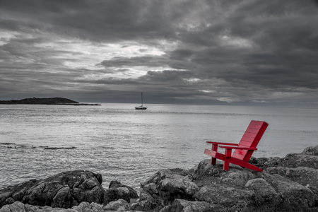 Red chair on ocean side rocks contrasts with the stormy black and white background.の写真素材