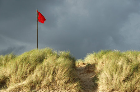 Red danger flag on a beach. Warning flag at the top of a grass-covered sand dune indicating unsafe...