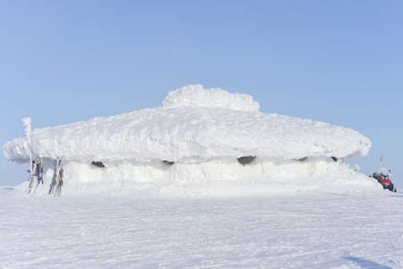 In the sunlight, this large, low lying building is covered in a very thick layer of bright white snow. Skis are rested against one end of the building.の写真素材