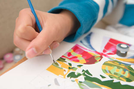 6 - Multicolored close up of painting by numbers. Young and bright female hand holds the bright blue paintbrush with detail as it paints greenの写真素材