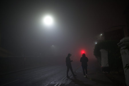 Hooded youths loiter on a street corner using mobile smartphones. Dark and foggy conditions with...