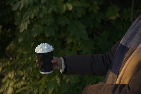 Young women in winter coat and scarf holds a takeaway beverage, hot chocolate, topped creamy marsh...