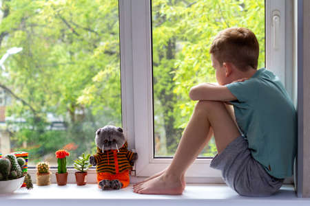Young boy sitting on windowsill and looking at lovely toy cat. Stay at home concept during pandemic of covid-19の写真素材