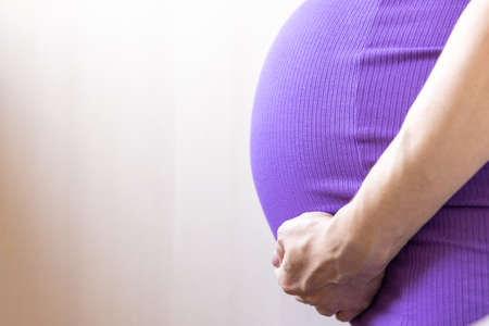 Unrecognizable Close up Pregnant women. Hands over tummy during last month of pregnancy. In violet color dress on light background. Hand on belly. Pregnancy, maternity, preparation concept.Copy space.の写真素材