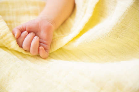Newborn baby hand. Close up little fist over yellow muslin blanket. With copy space. Happy family concept, daycare nursery. Childrens dayの写真素材