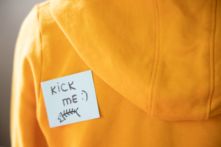 Boy in yellow hoodie with a sticky note on his back, saying kick me.の写真素材