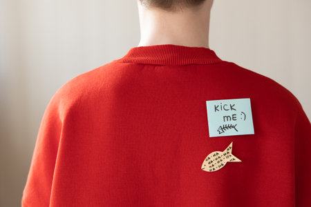Boy in red hoodie with a paper attached with tape to his back with the handwriting text kick me. Fools day jokeの写真素材