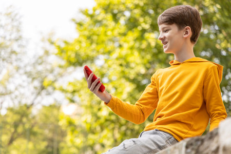 A teenager in a yellow hoodie, sitting in a park, smiles while looking at the screen of their...