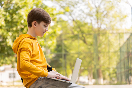 Thoughtful teenager boy resting. Holding and using a laptop for networking on a sunny day, outdoors.の写真素材