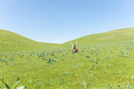 Back view of cheerful women sitting in the field with hands up, green field an blue sky view behind. Facelessの写真素材