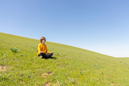 A young person sits peacefully on a green hillside. Tranquil and dreamy atmosphere. Peace and connection with natureの写真素材