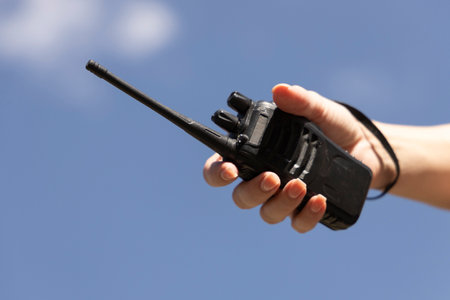 A portable walkie-talkie against a blue sky during a mountain adventure, held in a hand. Emphasizes communication, safety, and readiness in the wilderness. For tourism, extreme sports, teamwork, and outdoor technology themes. Dynamic composition with sunlight and fresh air.の写真素材