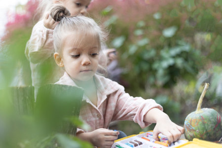 A little girl is painting a green pumpkin outdoors with colorful paints. Creativity, fun, and the charm of autumn craftsの写真素材