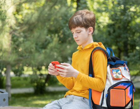 A teenage boy in a yellow hoodie sits in a park and looks at his smartphone. Green trees in the background create a relaxed outdoor settingの写真素材