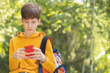 A teenage student in a yellow hoodie sits in a park and looks at his smartphone. Green trees in the background create a relaxed outdoor setting. Concept of education, online learning, and technologyの写真素材