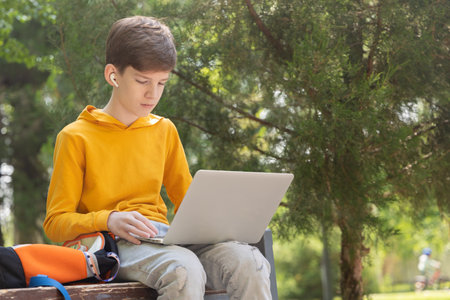 A teenage boy in a yellow hoodie sits in a park with a laptop, wearing wireless headphones. Green trees in the background create a natural summer setting. Side view.の写真素材