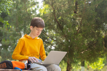 Thoughtful teenager boy working on laptop. Holding and using a laptop for networking on a sunny spring day, outdoors.の写真素材