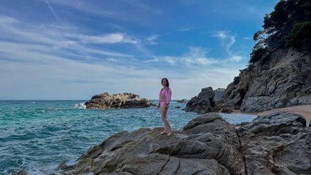 Young woman in a swimsuit staying on a rocky cliff at Playa Cala Sa Boadella in Lloret de Mar, Spain. Summer beach vibe with scenic seascape and natural textures. Banner for tourism promotion.の写真素材