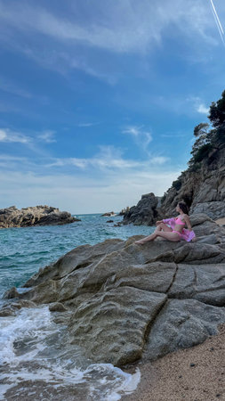 Summer photo of a slim woman in swimsuit sitting on rocky coast at Playa Cala Sa Boadella, Spain. Ideal for travel ads, hotel promotions, and tourism campaignsの写真素材