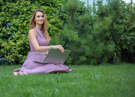 A girl in a dress on a green lawn in the park works at a laptopの写真素材