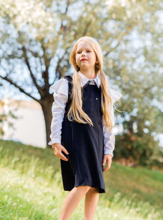 Little blonde girl with two ponytails, in a school uniform posing against a background of greenery and looking into the distance. Vertical photoの写真素材
