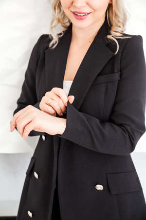 A blonde girl in a black suit stands against a white wall and straightens her sleeve. Vertical photoの写真素材