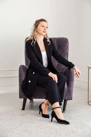 A blonde girl in a black suit and high-heeled shoes sits in a chair and poses at the camera. Vertical photoの写真素材