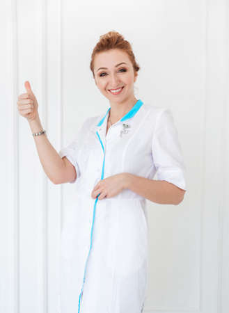 Beautiful young woman doctor posing in a white coat against a white wall background and showing class. Vertical photoの写真素材