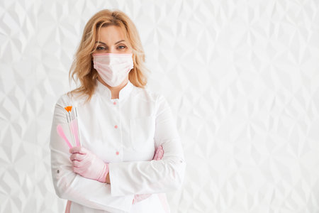 Beautiful young woman doctor beautician in a white uniform and a protective mask on her face posing...