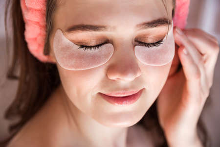 Beautiful young woman in a pink towel, with a cosmetic bandage on her head and with patches under her eyes. Girl sitting with closed eyes. Horizontal photoの写真素材