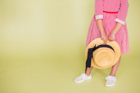 A girl in red and white sneakers holds a straw hat in her hands. Girl standing on a green...