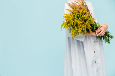 Girl in a light blue linen dress posing on a blue background with a bouquet of yellow wildflowers....