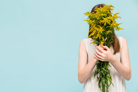 Girl in a white dress posing on a blue background covering her face with a bouquet of wildflowers....